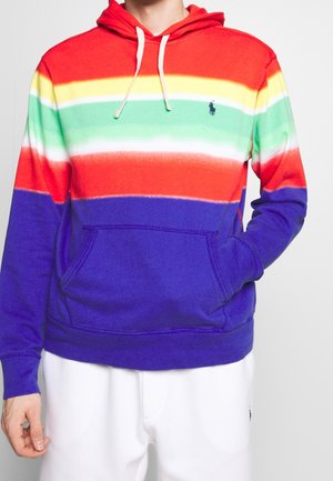 Mężczyzna w kolorowej bluzie z kapturem w poziome paski w kolorach czerwonym, żółtym, zielonym, pomarańczowym i niebieskim, z białymi sznureczkami, w zestawieniu z białymi spodniami.