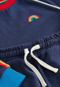 Outfit in cotone blu navy caratterizzato da un ricamo arcobaleno e un colletto rosso a coste. Include una vita elastica con cordino bianco e polsini colorati.