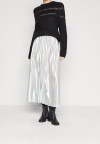 Maglione nero lavorato a maglia con dettagli in pizzo, abbinato a una gonna lunga plissettata argentata. Il modello indossa stivali neri con tacco alto.