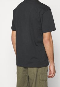 Camiseta de algodón negra con cuello redondo y mangas cortas, vista desde la espalda, combinada con pantalones cortos tipo cargo oliva.