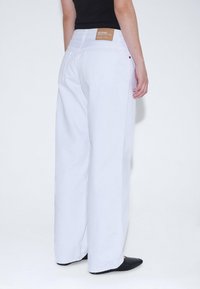 Pantalones vaqueros anchos blancos de algodón, con una etiqueta de cuero marrón en la parte posterior, dos bolsillos traseros y un corte recto. Zapatos negros visibles.