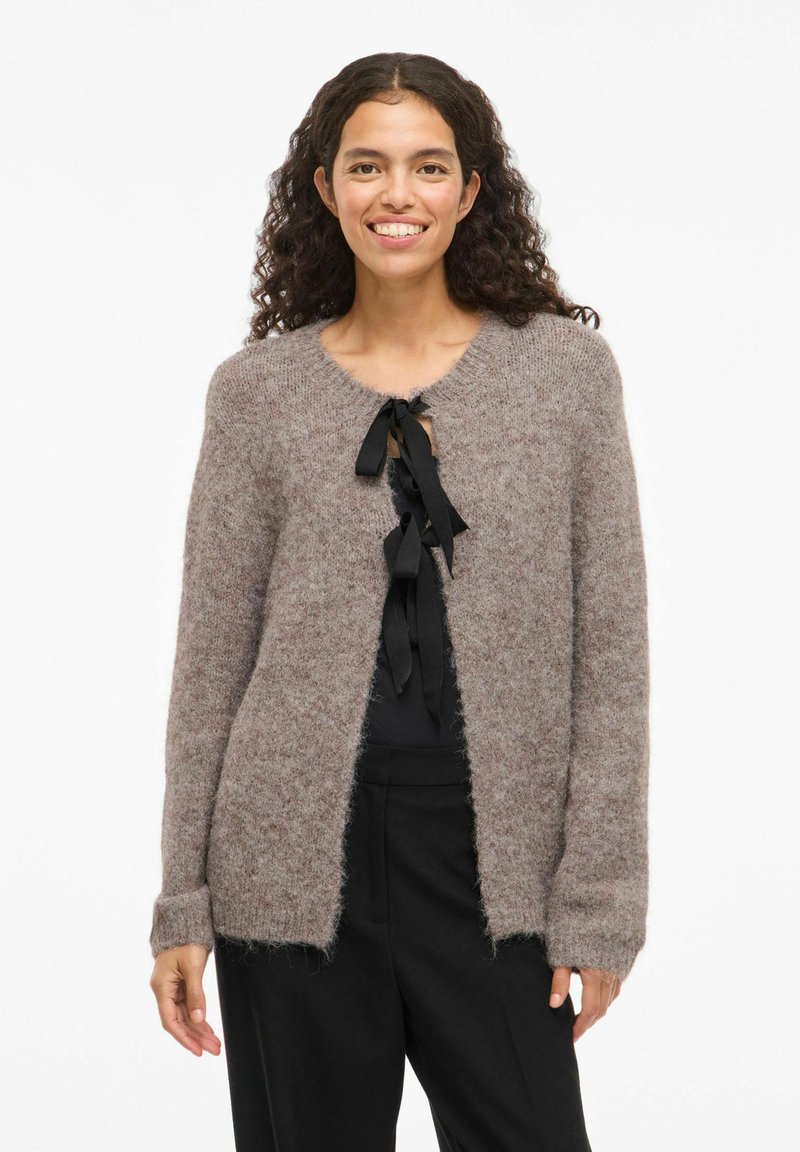 Gaiši brūns, pūkains adījuma kardigans ar apaļu kaklu, atvērtu priekšpusi un melnu lentu kā aizdari. Garas piedurknes ar ribotām aprocēm.