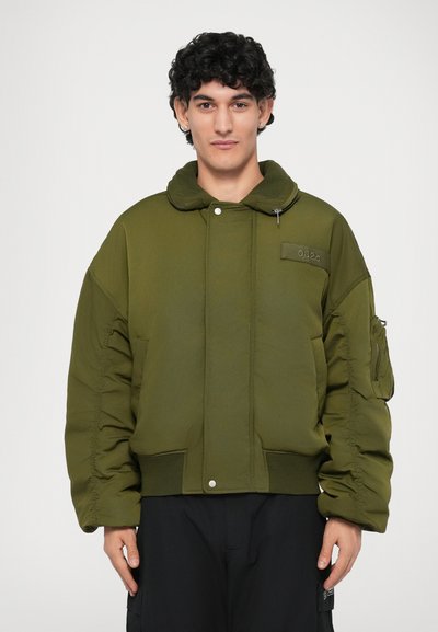 Olijfgroene pufferjas met een ritssluiting capuchon, zijzakken en elastische manchetten. Beschikt over stikseldetails en een logopatch op de borst.