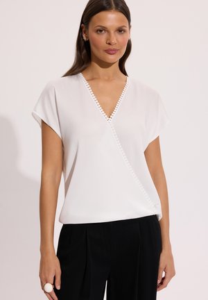 Kobieta w białej bluzce z dekoltem w serek z ozdobnym falbankowym wykończeniem i krótkimi rękawami, w połączeniu z czarnymi plisowanymi spodniami na jednolitym tle.