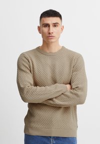 Beige neulepaita, jossa on teksturoitu pesäkuvio, ribba-ahtaat kaulus, hihansuut ja helma. Malli seisoo kädet ristissä, päällään siniset farkut.