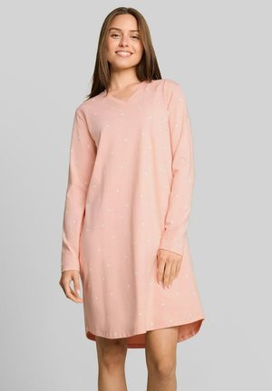 Jonge vrouw lacht, draagt een pastelroze jurk met lange mouwen en kleine witte stippen, staand tegen een effen lichtgrijze achtergrond.