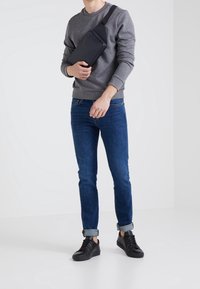Homme en sweat-shirt gris, jean bleu et baskets noires tenant un sac bandoulière noir sur un fond blanc uni.