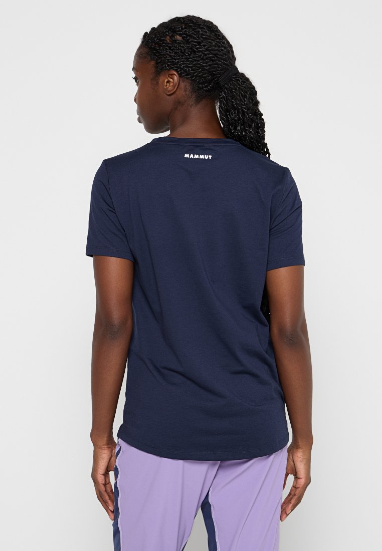 Vrouw met gevlochten haar, gekleed in een marineblauwe Mammut t-shirt en lavendel sportbroek, van achteren gezien tegen een effen achtergrond.