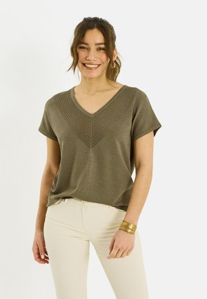 Mujer sonriente con cabello castaño recogido en una coleta que lleva una blusa de cuello en V texturizada de color verde oliva, pantalones crema y pulseras doradas sobre un fondo blanco.