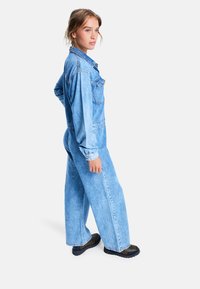 Jumpsuit de denim en azul claro con un corte relajado, mangas largas, bolsillos frontales y collar con botones, combinado con zapatos negros gruesos.