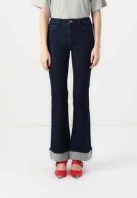 Jean flare bleu foncé avec taille haute, présentant un ourlet retroussé en denim clair, associés à des chaussures pointues rouges.