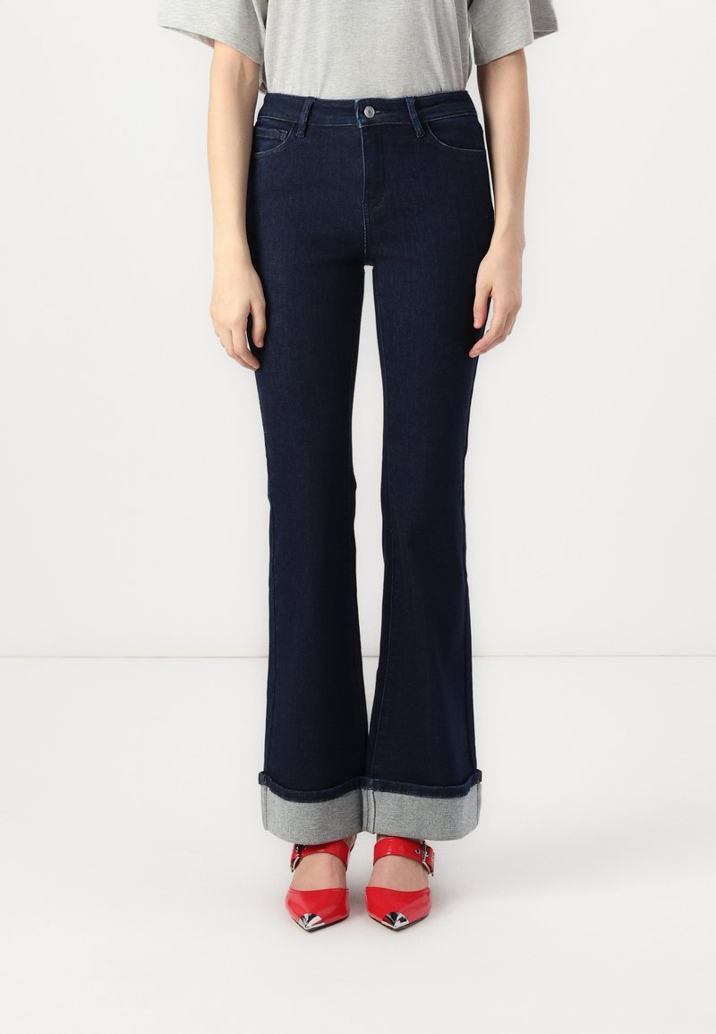 Jean flare bleu foncé avec taille haute, présentant un ourlet retroussé en denim clair, associés à des chaussures pointues rouges.