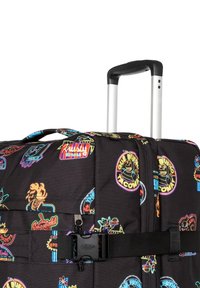 Rollen-Duffelbag aus schwarzem Stoff mit bunten, cartoonartigen Aufnähern und Logos, ausgestattet mit einem ausziehbaren Griff und einem Schnallenverschluss.