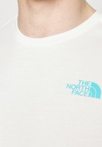 Camiseta de algodón blanca con un logo en azul claro de "The North Face" en el pecho izquierdo. Cuello redondo y textura suave.