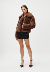 Καφέ ψεύτικο γούνινο μπουφάν με φαρδύ γιακά, μακριά μανίκια και κοντό σχέδιο, συνδυασμένο με ένα μαύρο εφαρμοστό φόρεμα και μαύρα μυτερά τακούνια.