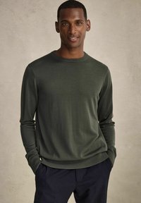 Grön stickad tröja med rund halsringning och långa ärmar, ribbade ärmslut och nederkant, kombinerad med mörka byxor, mot en neutral bakgrund.