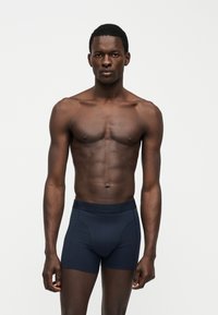 Αρσενικό μοντέλο με σκούρο δέρμα, στέκεται μπροστά σε απλό φόντο φορώντας σκούρα μπλε εφαρμοστά boxer, κοιτάζοντας προς τα εμπρός με χαλαρά χέρια.