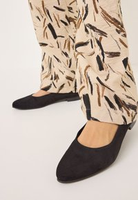Ballerines en suédine noire au design à bout pointu. Portées avec un pantalon beige et marron à motifs présentant des designs de coups de pinceau abstraits.