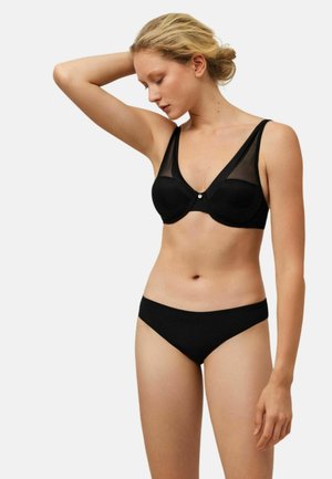 Mujer con cabello rubio, con un sujetador negro con paneles translúcidos y ropa interior negra a juego, posando con un brazo levantado detrás de la cabeza.