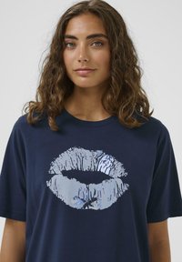 Navyblaues Baumwoll-T-Shirt mit einem großen, glänzenden, silbernen Lippenabdruck-Design auf der Vorderseite, einem Rundhalsausschnitt und kurzen Ärmeln.