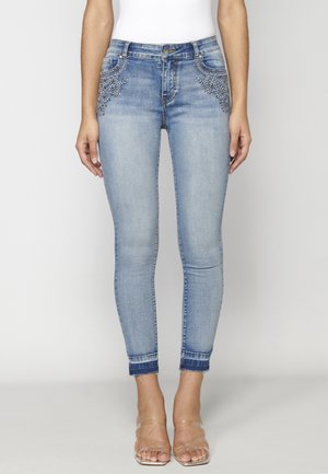 Frau trägt hellblaue Skinny-Jeans mit bestickten Blumendetails in der Nähe der Vordertaschen und transparente Sandalen mit Absatz.