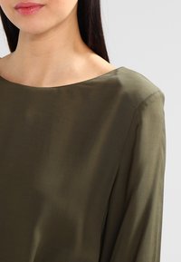 Olivově zelený top s lodičkovým výstřihem, hladkým povrchem a dlouhými rukávy, s jednoduchým a elegantním designem bez ozdob.