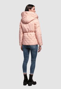 Chaqueta acolchada rosa con una gran capucha, patrón de acolchado en diamante y cintura ajustada. Usada sobre jeans azules y botas negras de tobillo.