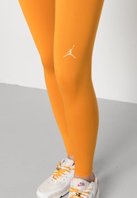Leggings arancioni realizzati in tessuto elasticizzato, con un logo Jumpman bianco sulla gamba, abbinati a sneakers bianche con lacci arancioni.