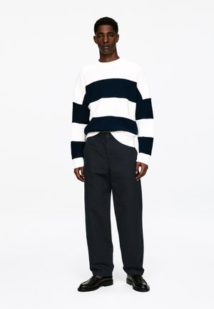 Biały i granatowy sweter w paski o teksturowanym splocie, zestawiony z ciemnogranatowymi spodniami i czarnymi skórzanymi butami.