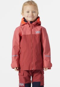 Chaqueta impermeable roja con detalles en naranja, que cuenta con un frente con cremallera, capucha y un logo gris. Combinada con pantalones a juego, también en rojo y azul marino.