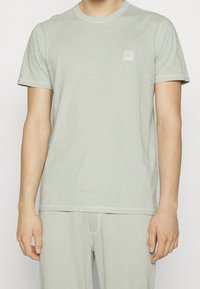 Ljusgrön bomulls T-shirt med korta ärmar, rundad halsringning och en liten fyrkantig logotyp på vänster bröst. Mjuk textur och minimalistisk design.