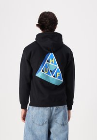 Schwarzer Hoodie mit einem blauen und türkisfarbenen dreieckigen Design auf dem Rücken, mit gelben Buchstaben "H", "U" und "F". Weicher Stoff und entspannte Passform.