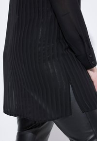 Zwarte geplisseerde blouse met een doorschijnende textuur, lange mouwen, knopen bij de manchetten en zijsplitten, gecombineerd met slanke zwarte leren broek.