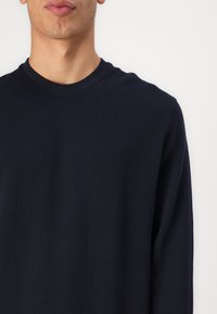 Camisa de cuello redondo de manga larga en azul marino, hecha de un tejido texturizado con un sutil patrón de espiga, que presenta un escote clásico y mangas ajustadas.