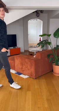 Narancssárga bordázott szövet sarokkanapé, fa padló, nagy zöld növény, lapos képernyős TV és kör alakú üveg kávézóasztal egy világos szobában.