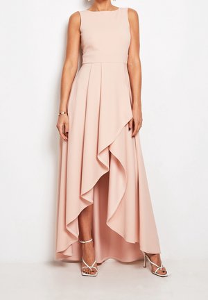 Femme portant une robe rose blush sans manches, courte devant et longue derrière, avec un ourlet asymétrique à volants et des sandales blanches à talons hauts à brides.