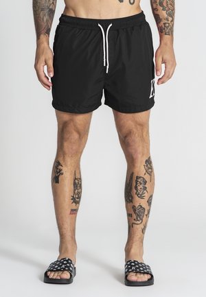 Uomo con pantaloncini neri con coulisse bianca e sandali neri, che mostra braccia e gambe tatuate su sfondo neutro.