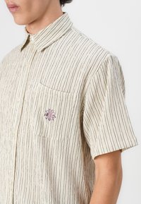 Kortermet skjorte med knappestilling i kremfarge med tynne vertikale striper, som har en brystlomme og en brodert logo i rødt og svart.