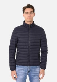 Giacca puffer blu navy con alto colletto, texture trapuntata, zip frontale e tasche laterali. Indossata sopra un paio di jeans blu.