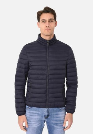 Giacca puffer blu navy con alto colletto, texture trapuntata, zip frontale e tasche laterali. Indossata sopra un paio di jeans blu.