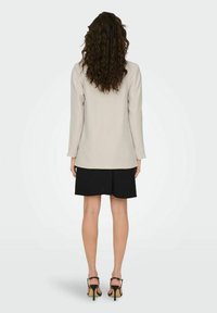 Beige op maat gemaakte blazer met lange mouwen, gecombineerd met zwarte shorts en zwarte hoge hakken. Krullend haar zichtbaar van achteren.