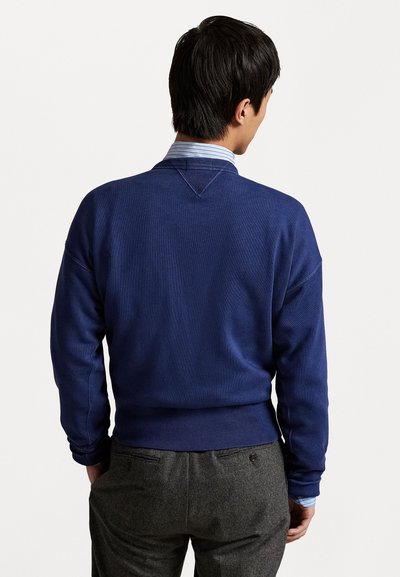 Un sweatshirt bleu marine à col rond avec des poignets et un ourlet côtelés. Il présente un tissu texturé, porté par-dessus une chemise rayée bleu clair et blanche.