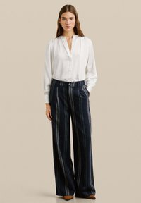 Chemise blanche à manches longues avec épaules froncées, associée à un pantalon bleu marine taille haute et large avec des rayures verticales et un détail de bouton.