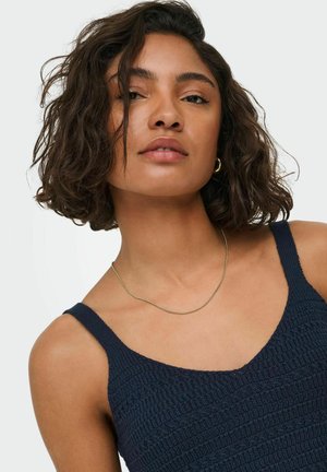 Young woman with short curly dark hair wearing a navy blue textured tank top, gold hoop earring, and thin gold necklace.
