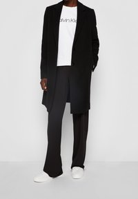 Cappotto lungo nero, felpa bianca con logo, pantaloni neri e sneakers bianche. Il cappotto ha una vestibilità su misura e un design elegante.