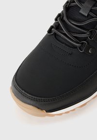 Black sneaker with textured upper, featuring dark eyelets and laces. Light brown rubber sole with white accent. Minimalist design.