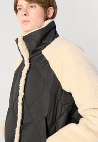 Μαύρη μονωμένη τζάκετ με κρεμ φωτισμένες λεπτομέρειες στο γιακά και στους ώμους; καπιτονέ υφή; κλείσιμο μπροστά με κουμπιά.