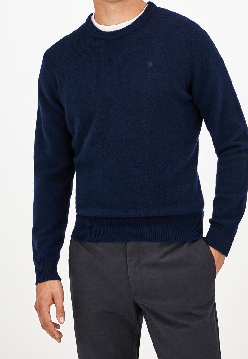 Homme portant un pull à col rond bleu marine sur une chemise blanche et un pantalon gris foncé, debout avec une main dans la poche, sur fond blanc.