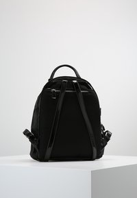 Mustassa verkkomateriaalista ja kiiltävästä vinyylistä valmistettu reppu, jossa on säädettävät hihnat, yläkahva ja vetoketjukiinnitys. Yksinkertainen, rakenteellinen muotoilu.