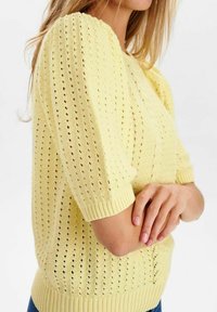 Gul stickad tröja med korta ärmar, med ett strukturerat mönster av öppna hål, och ribbade kanter vid muddarna och fållen.